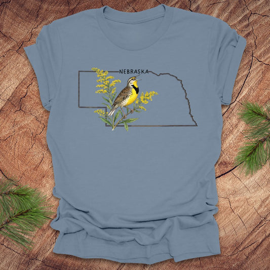 Steel blue t-shirt with the outline of the state of Nebraska and a  graphic of a meadowlark and goldenrod inside on a wooden surface