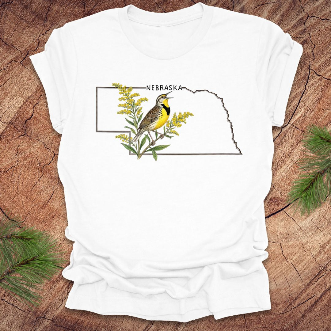 White t-shirt with the outline of the state of Nebraska and a  graphic of a meadowlark and goldenrod inside on a wooden surface