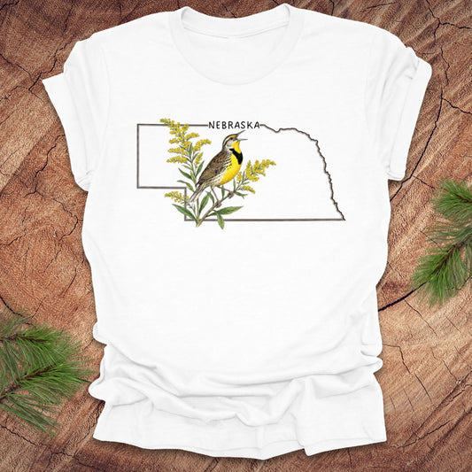 White t-shirt with the outline of the state of Nebraska and a  graphic of a meadowlark and goldenrod inside on a wooden surface