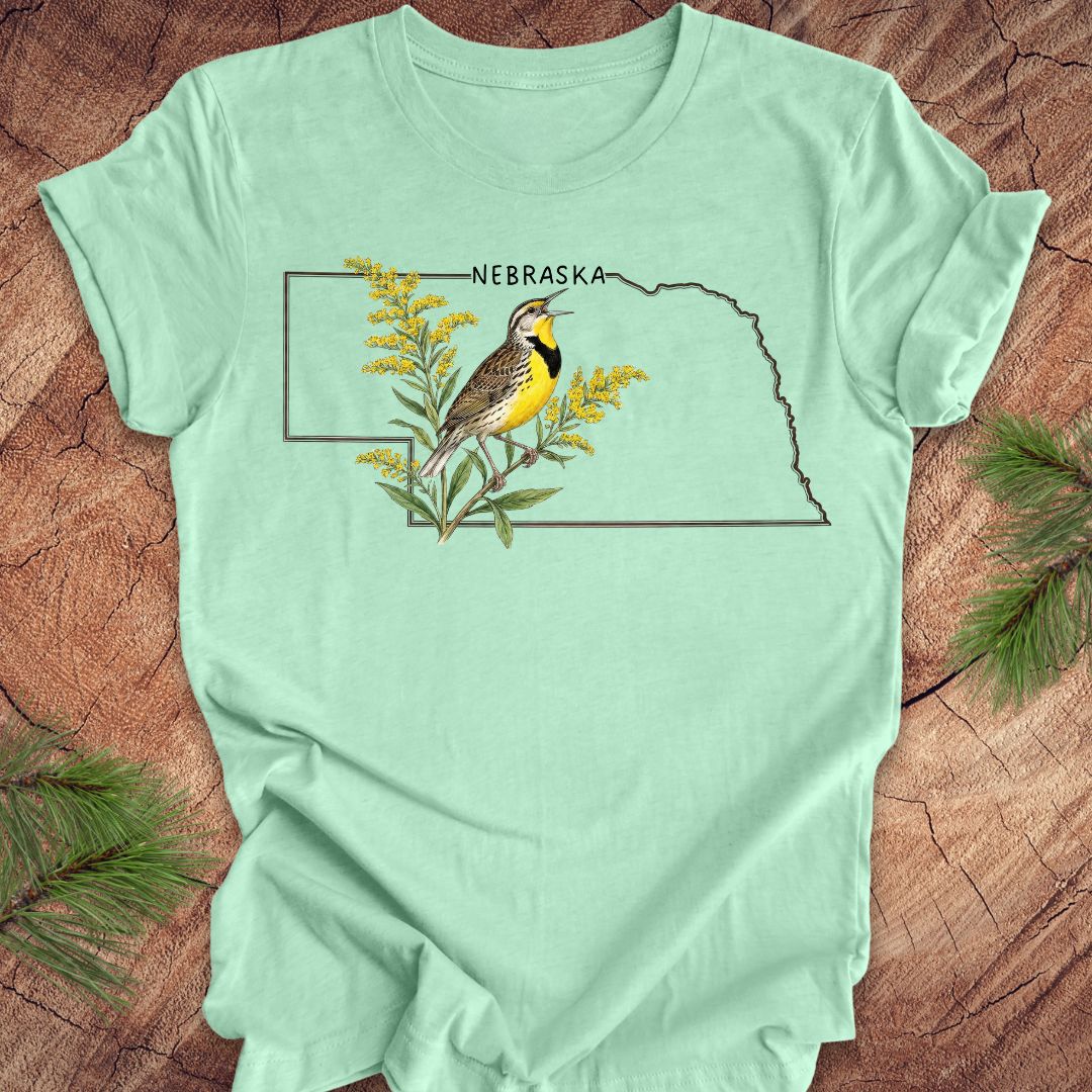 Mint green t-shirt with the outline of the state of Nebraska and a  graphic of a meadowlark and goldenrod inside on a wooden surface