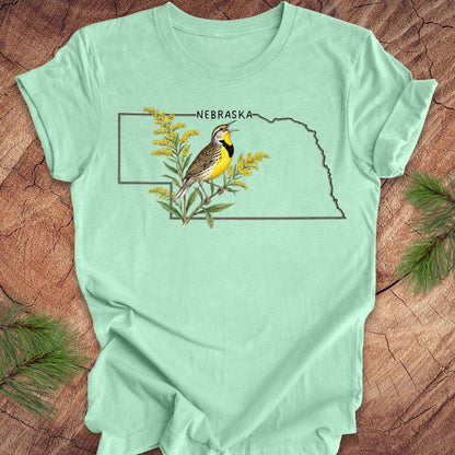 Mint green t-shirt with the outline of the state of Nebraska and a  graphic of a meadowlark and goldenrod inside on a wooden surface