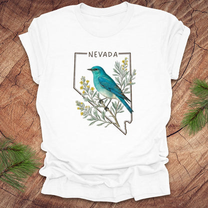 White t-shirt with Nevada state design featuring a mountain bluebird and sagebrush on a wooden surface.