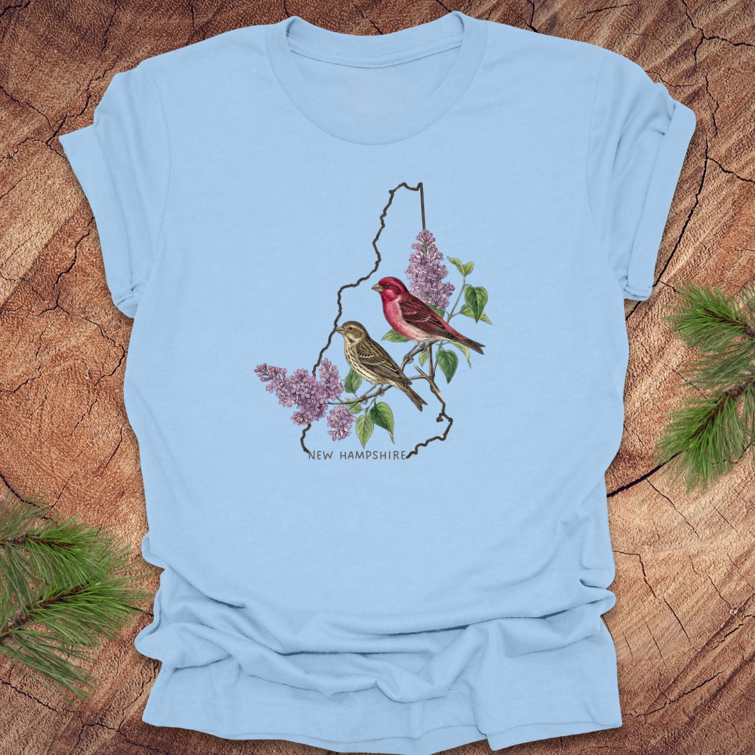 Baby blue t-shirt with New Hampshire design featuring purple finches and purple lilac on a wooden surface.