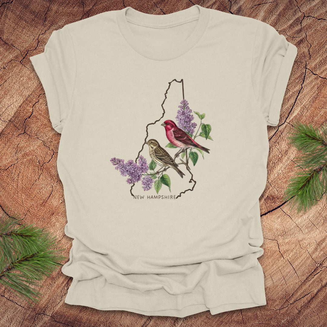 Beige t-shirt with New Hampshire design featuring purple finches and purple lilac on a wooden surface.