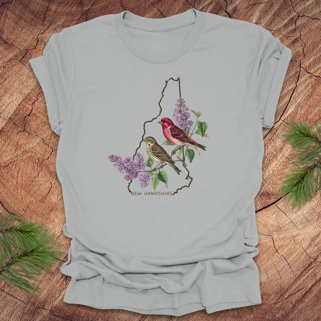 Silver t-shirt with New Hampshire design featuring purple finches and purple lilac on a wooden surface.