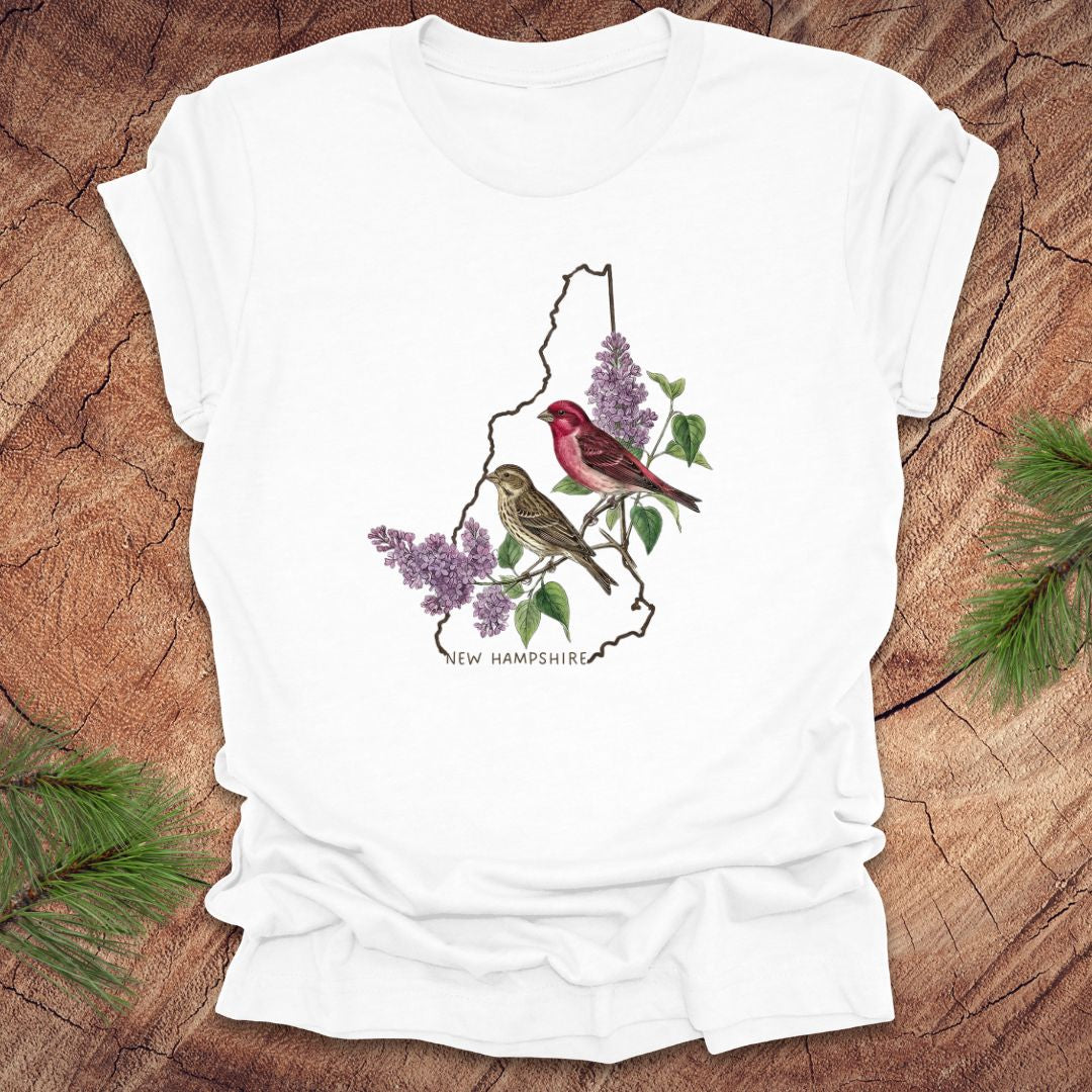 White t-shirt with New Hampshire design featuring purple finches and purple lilac on a wooden surface.