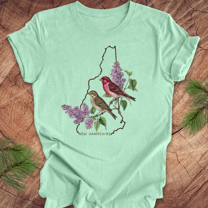 Mint green t-shirt with New Hampshire design featuring purple finches and purple lilac on a wooden surface.