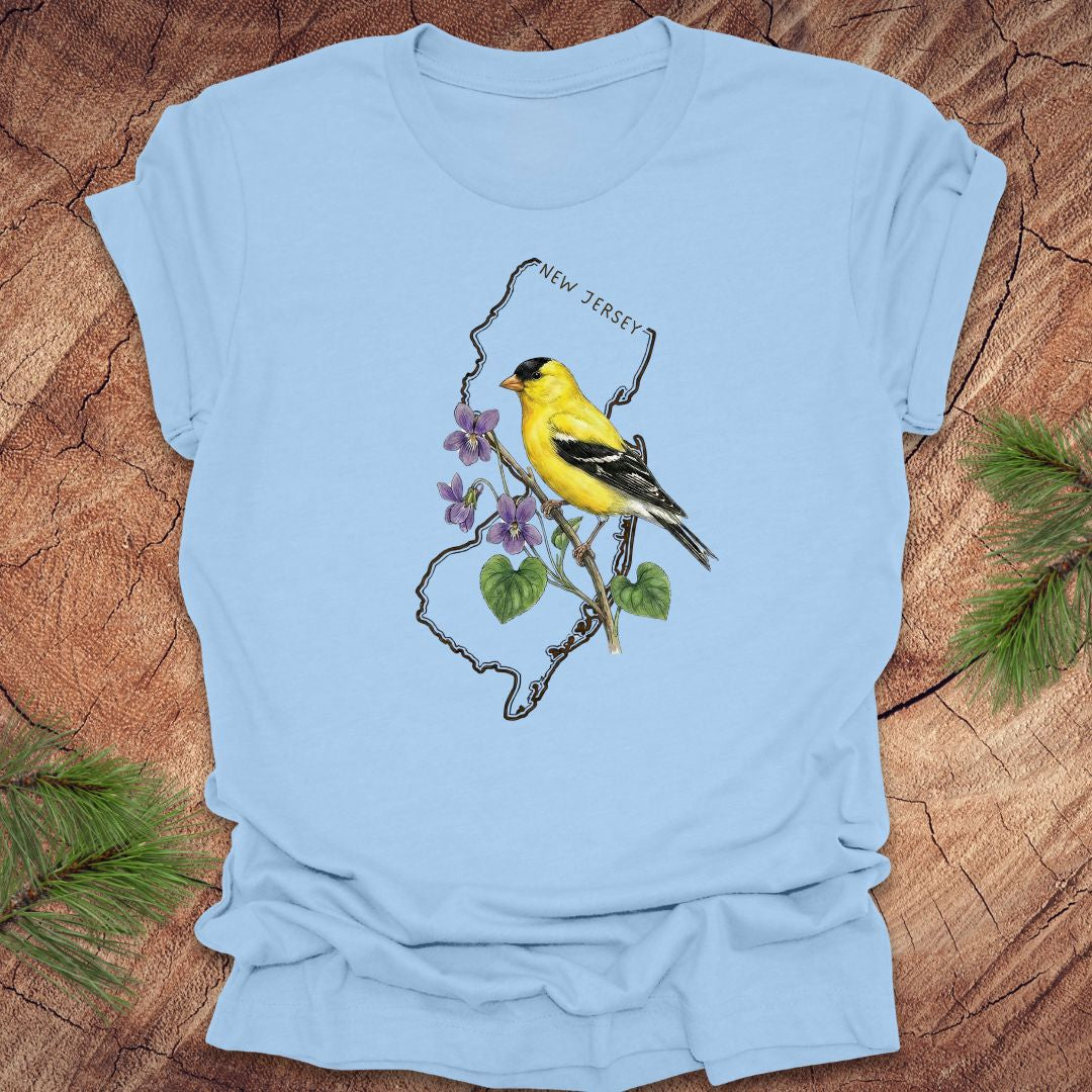 Baby blue t-shirt with a goldfinch and violets in a New Jersey state outline design on a wooden surface