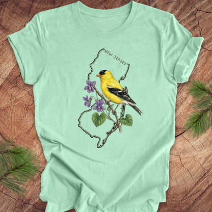 Mint green t-shirt with a goldfinch and violets in a New Jersey state outline design on a wooden surface