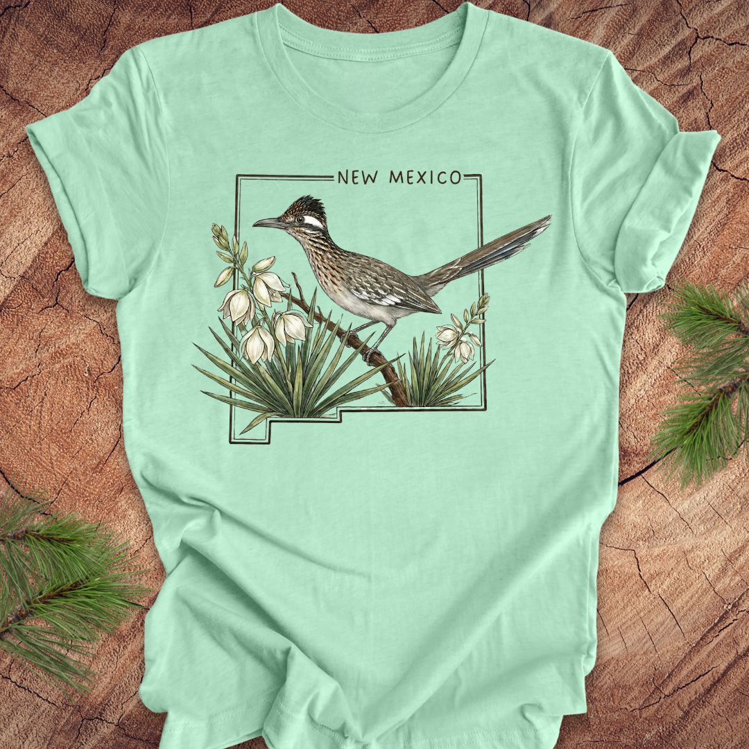 Mint green t-shirt with outline of state of New Mexico with state bird, roadrunner and flower yucca, on a wooden surface.