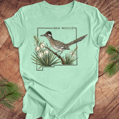Mint green t-shirt with outline of state of New Mexico with state bird, roadrunner and flower yucca, on a wooden surface.