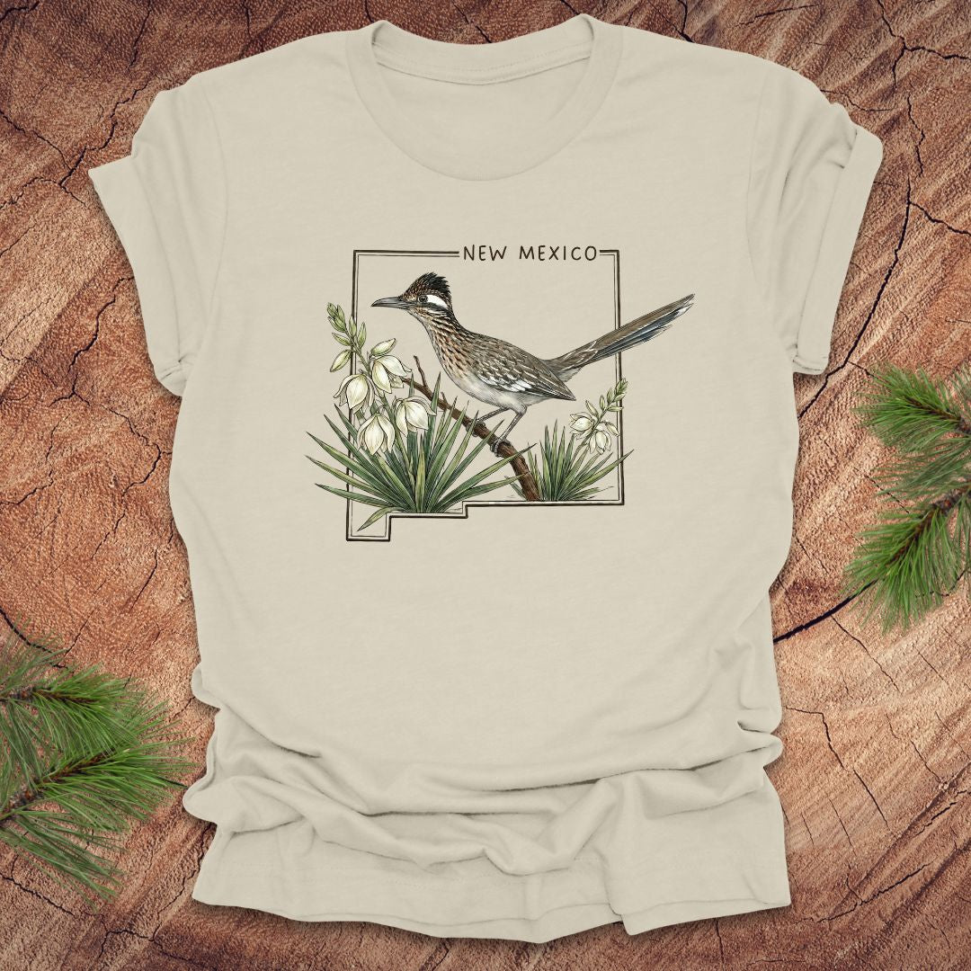 Beige t-shirt with outline of state of New Mexico with state bird, roadrunner and flower yucca, on a wooden surface.