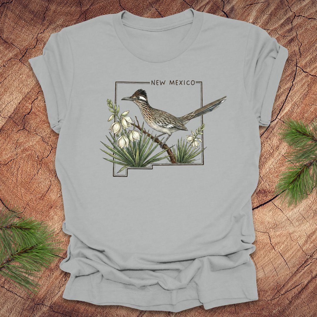Silver t-shirt with outline of state of New Mexico with state bird, roadrunner and flower yucca, on a wooden surface.