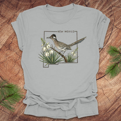 Silver t-shirt with outline of state of New Mexico with state bird, roadrunner and flower yucca, on a wooden surface.
