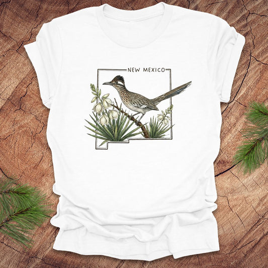 White t-shirt with outline of state of New Mexico with state bird, roadrunner and flower yucca, on a wooden surface.