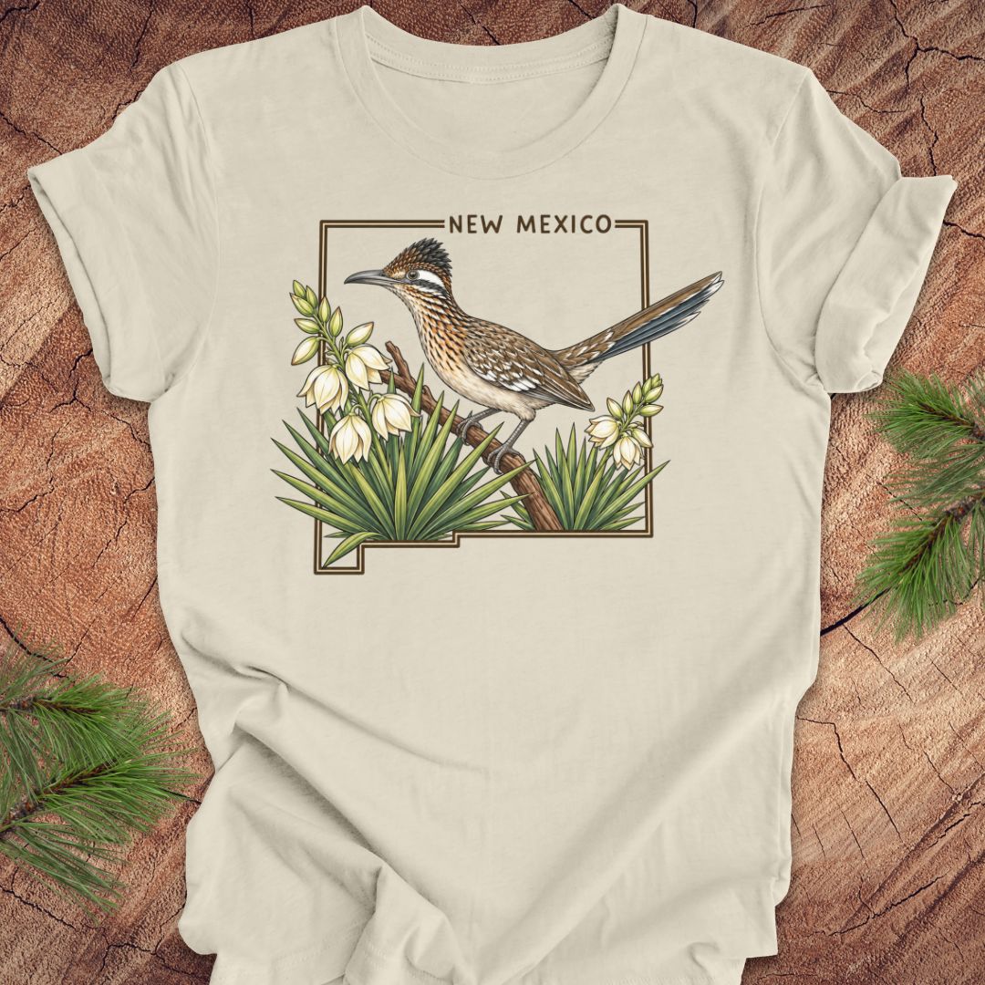 An off-white t-shirt with a 'New Mexico' roadrunner bird graphic, laid flat on a rustic wood background with pine branches.