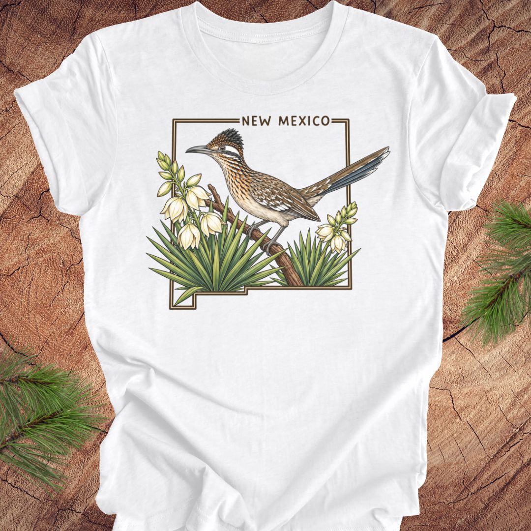 A white short-sleeve crewneck t-shirt laid flat on a rustic wood surface, featuring a detailed graphic of a roadrunner perched on a branch with white flowers and green agaves, framed by the text 'NEW MEXICO'.