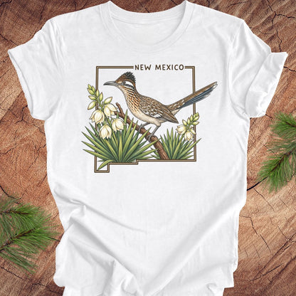 A white short-sleeve crewneck t-shirt laid flat on a rustic wood surface, featuring a detailed graphic of a roadrunner perched on a branch with white flowers and green agaves, framed by the text 'NEW MEXICO'.
