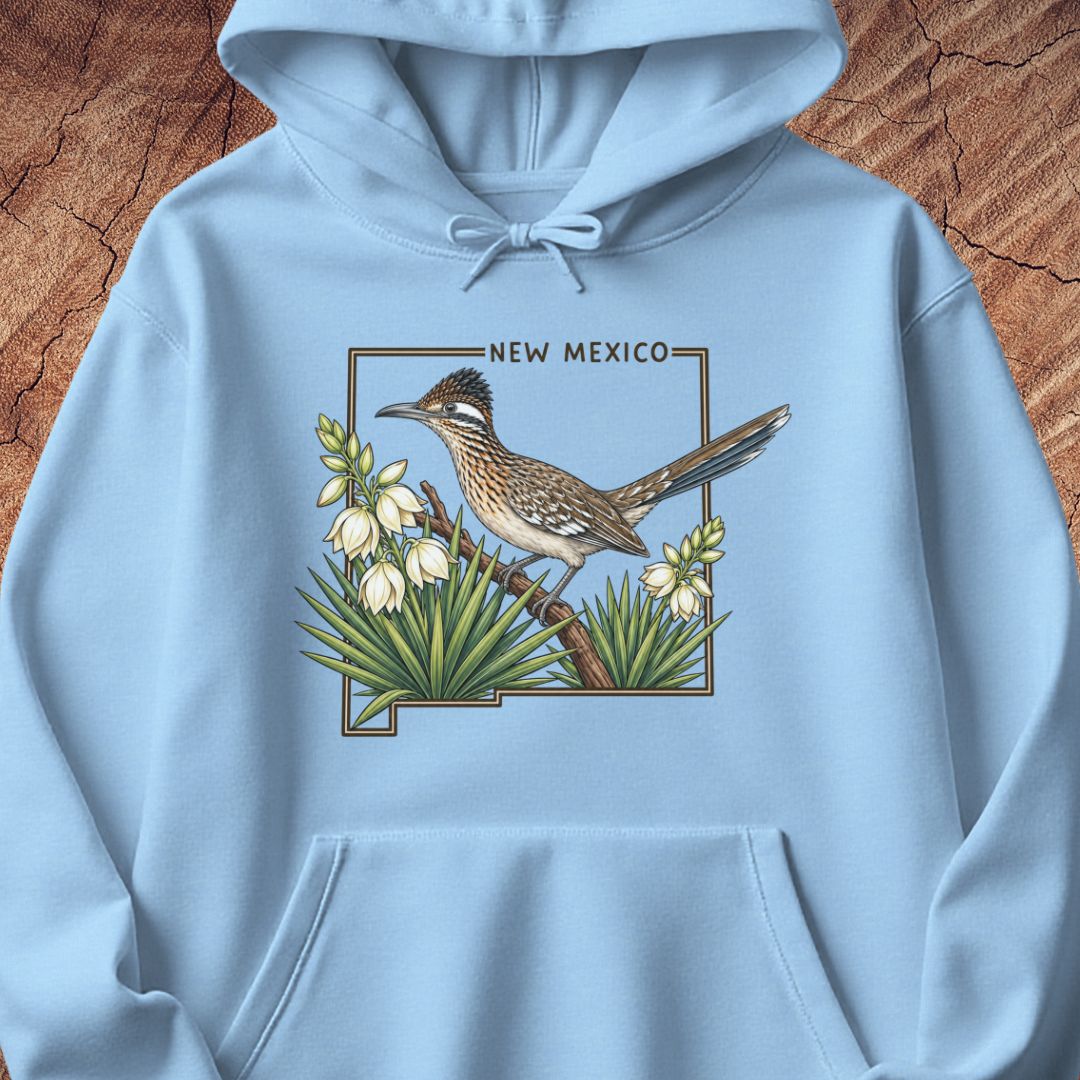A light blue pullover hoodie laid flat on a textured brown wood background, featuring a central graphic of a roadrunner bird perched among white flowers with the words 'NEW MEXICO' above it.
