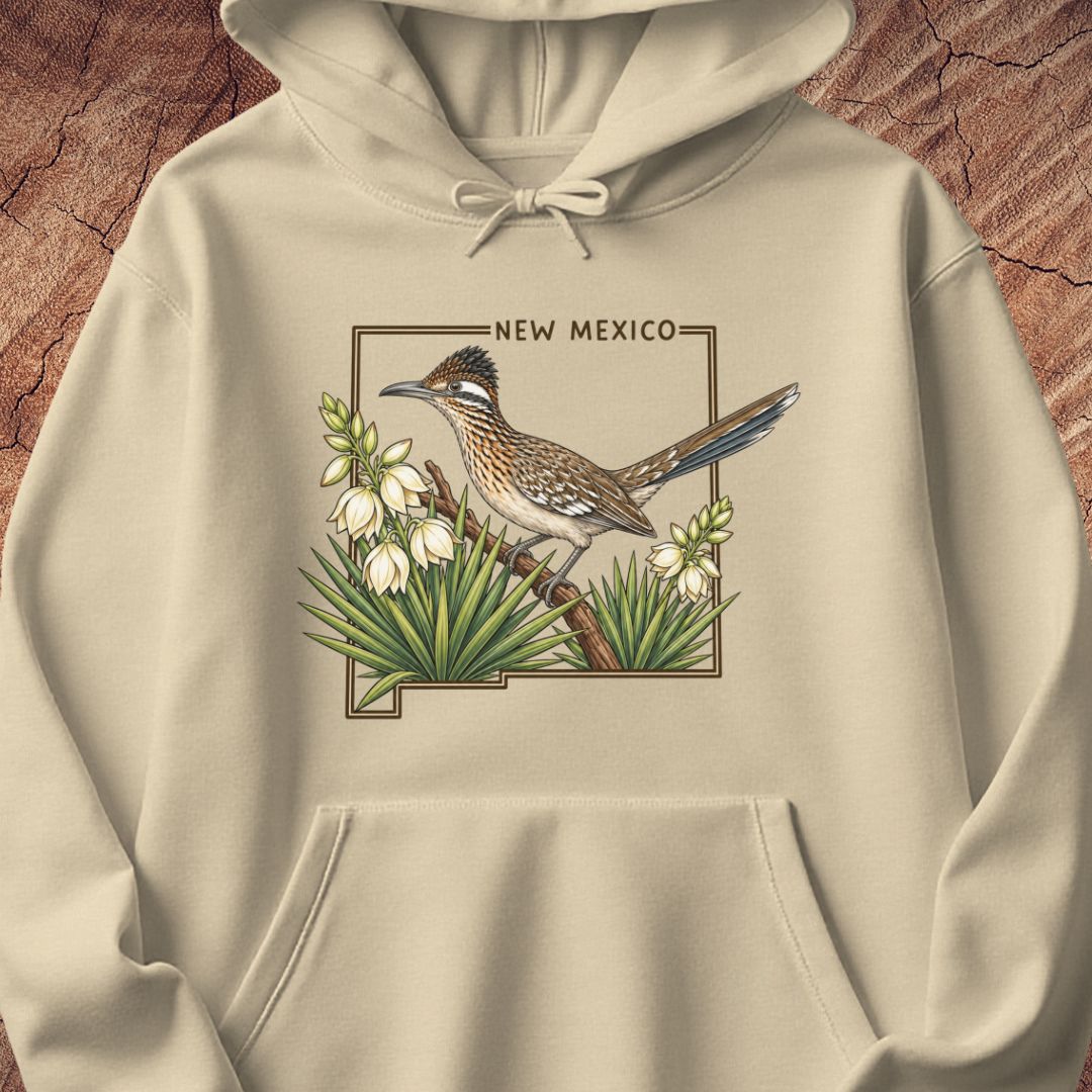 A beige pullover hoodie featuring a detailed graphic of a roadrunner bird perched on a branch with white flowers and green yucca plants, framed by the text 'NEW MEXICO'. The hoodie is displayed flat against a textured brown wood background.