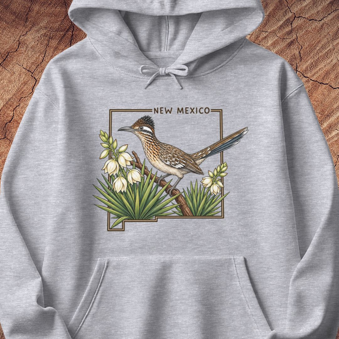 A light heather gray pullover hoodie featuring a detailed graphic print of a roadrunner bird perched among white flowers, with the text 'NEW MEXICO' above. The hoodie is displayed flat against a rustic wood background.