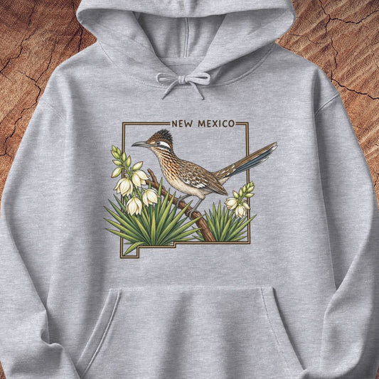 A light heather gray pullover hoodie featuring a detailed graphic print of a roadrunner bird perched among white flowers, with the text 'NEW MEXICO' above. The hoodie is displayed flat against a rustic wood background.