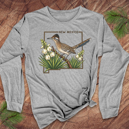 A heather gray long-sleeve t-shirt with a 'New Mexico' roadrunner bird graphic, styled flat on a rustic wood background with pine needles.