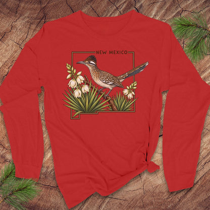 A red long-sleeve t-shirt with a graphic of a roadrunner bird, white flowers, and the outline of New Mexico, displayed flat on a rustic wood background with pine needles.