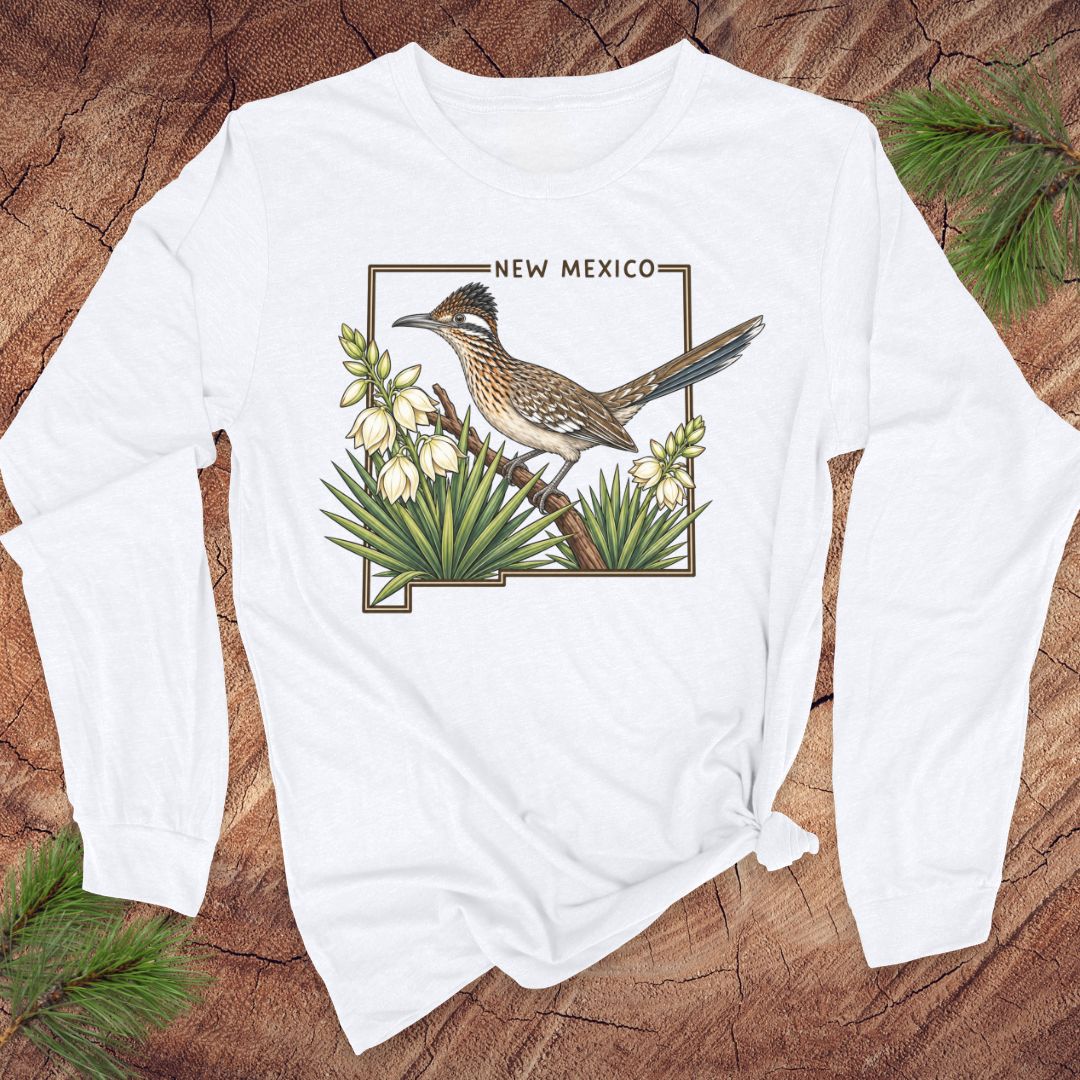 A white long-sleeve t-shirt with a graphic of a roadrunner bird and 'NEW MEXICO' text, laid flat on a rustic wood background with pine branches.