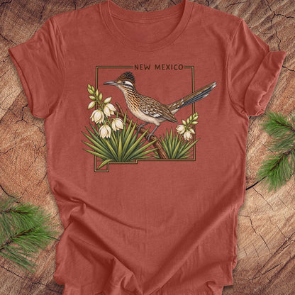A terracotta-colored short-sleeve t-shirt featuring a graphic of a roadrunner bird perched on yucca plants with the text 'NEW MEXICO' above, laid flat on a rustic wood background with pine branches.