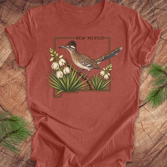 A terracotta-colored short-sleeve t-shirt featuring a graphic of a roadrunner bird perched on yucca plants with the text 'NEW MEXICO' above, laid flat on a rustic wood background with pine branches.