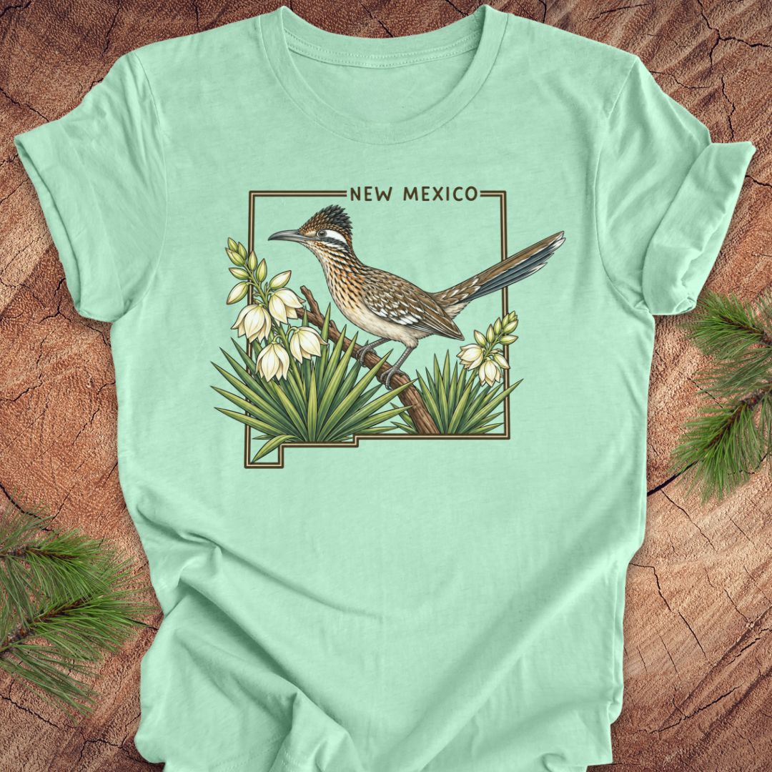 A mint green short-sleeve t-shirt laid flat on a rustic wood surface, featuring a vintage-style illustration of a roadrunner bird perched among white flowers with the words 'NEW MEXICO' above it.