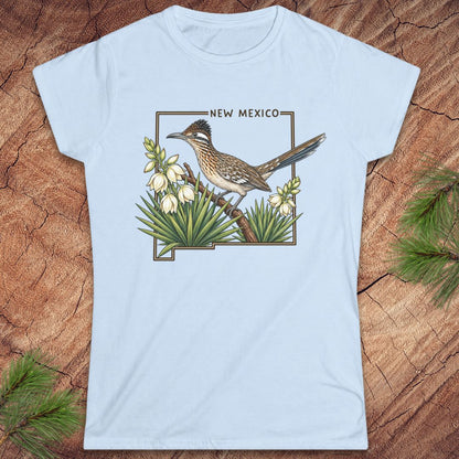 A light blue short-sleeve t-shirt with a graphic of a roadrunner bird and white flowers inside the outline of New Mexico, displayed flat on a rustic wood background with pine needles.