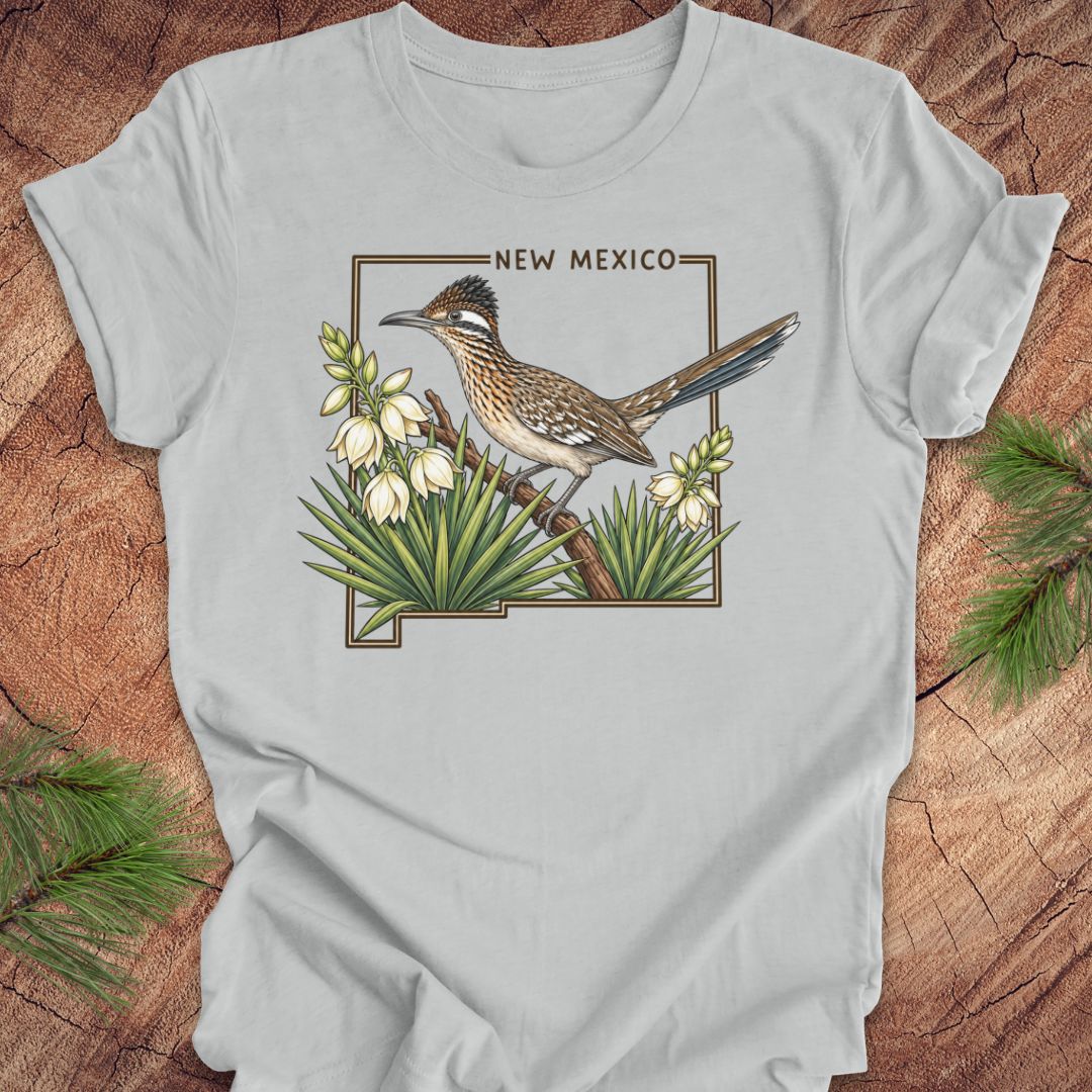 A light gray crewneck t-shirt featuring a vintage-style graphic of a roadrunner bird perched on a branch with white flowers and green leaves, framed by the text 'NEW MEXICO'. The t-shirt is laid flat on a rustic wood background with pine needles in the corners.