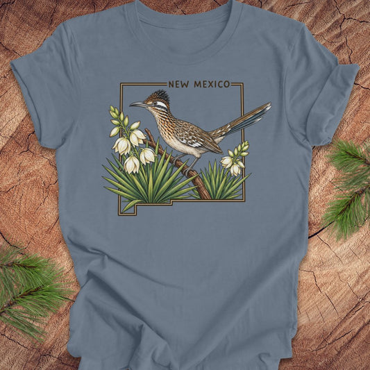 A slate blue short-sleeve t-shirt with a detailed graphic of a roadrunner bird perched on a branch with white flowers, framed by the text 'NEW MEXICO' above. The shirt is laid flat on a rustic wood background with pine needles in the corners.