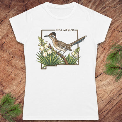 A white short-sleeve crewneck t-shirt featuring a printed graphic of a roadrunner bird perched on a branch with white flowers and green agave plants, framed by the outline of New Mexico with the text 'NEW MEXICO' above. The shirt is displayed flat on a rustic wood background with pine needles in the corners.
