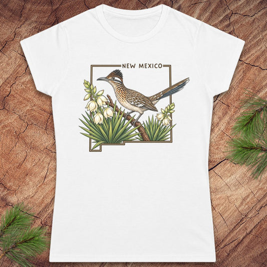 A white short-sleeve crewneck t-shirt featuring a printed graphic of a roadrunner bird perched on a branch with white flowers and green agave plants, framed by the outline of New Mexico with the text 'NEW MEXICO' above. The shirt is displayed flat on a rustic wood background with pine needles in the corners.