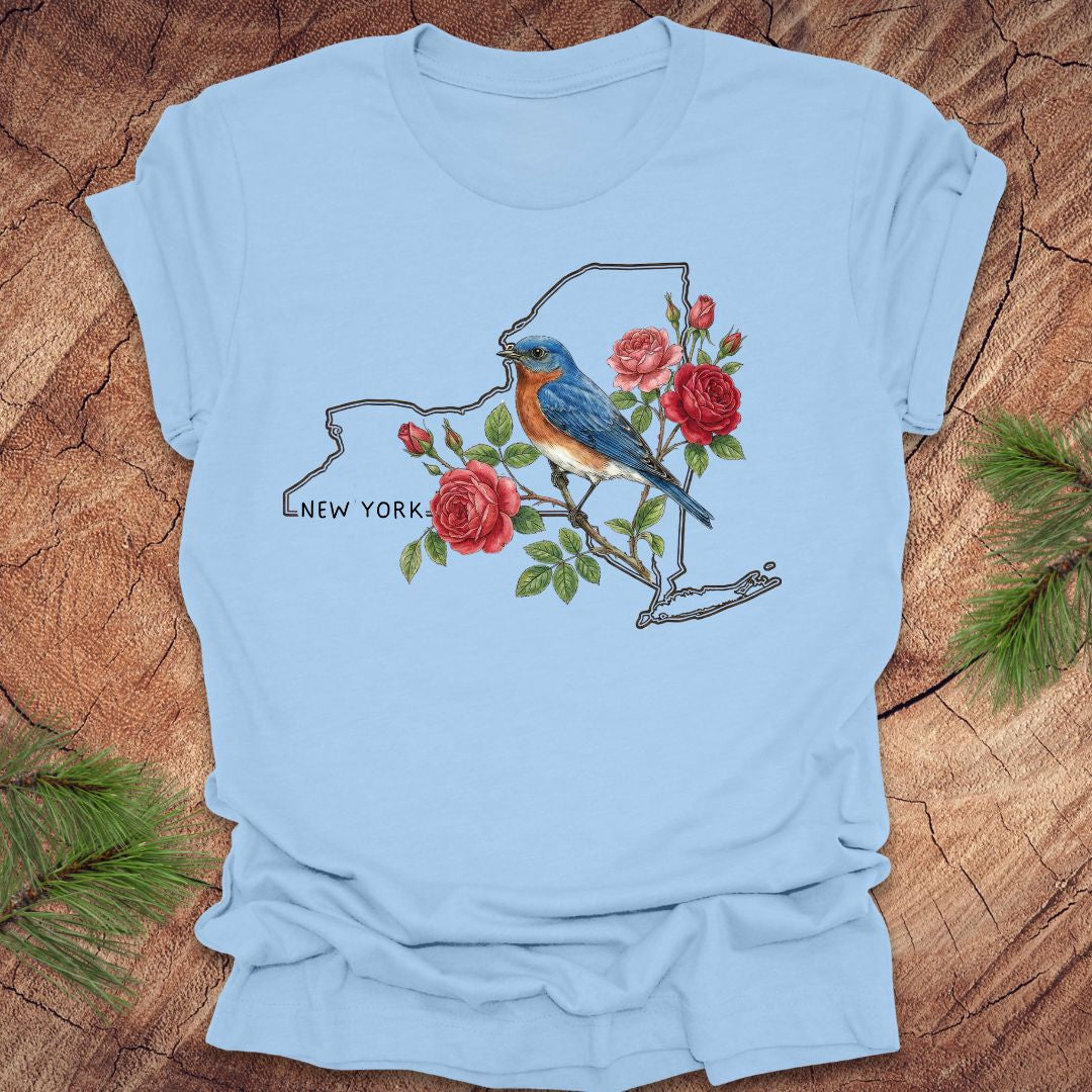 Baby blue t-shirt with a New York state outline featuring a bluebird and roses on a wooden surface.