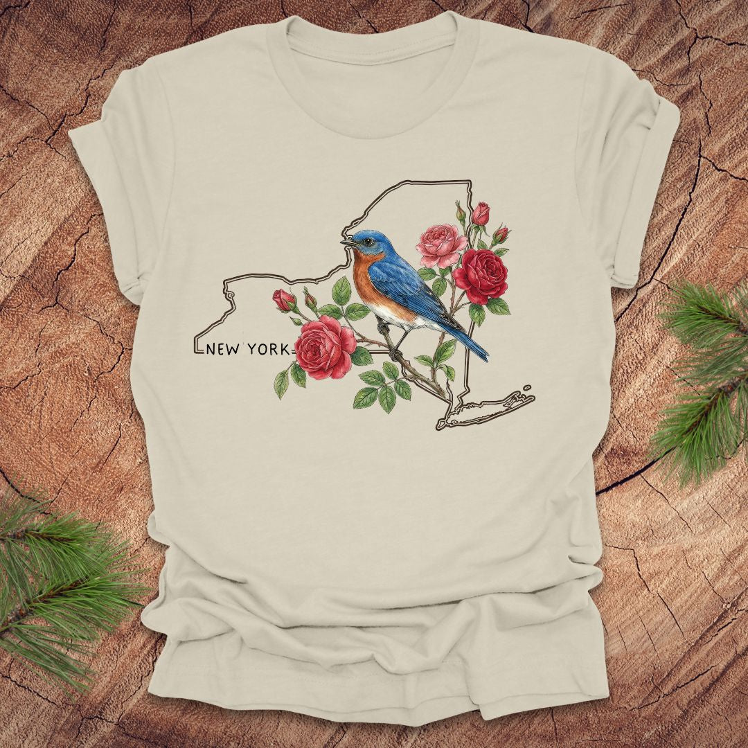 Beige t-shirt with a New York state outline featuring a bluebird and roses on a wooden surface.