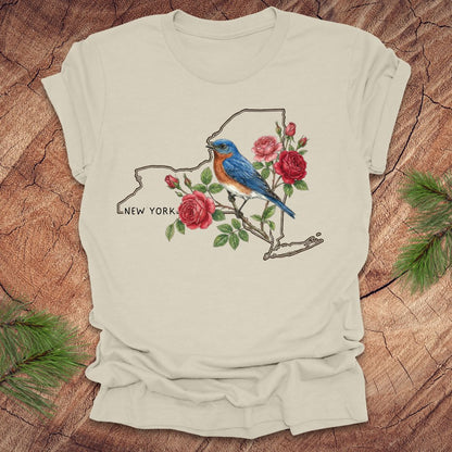 Beige t-shirt with a New York state outline featuring a bluebird and roses on a wooden surface.