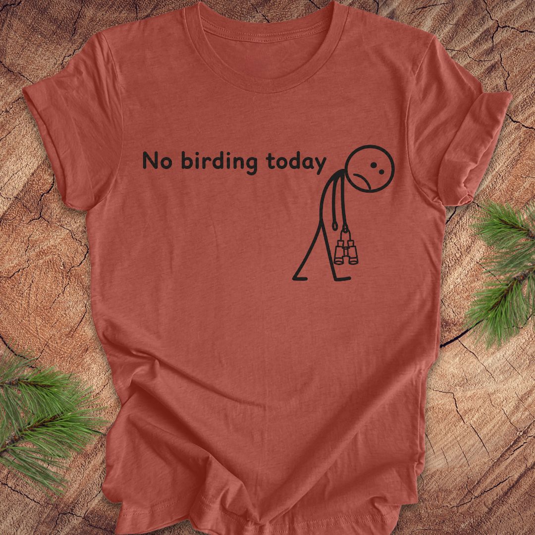 A terracotta-colored short-sleeve t-shirt with a black graphic print that reads 'No birding today' next to a sad stick figure holding binoculars. The shirt is laid flat on a rustic wood background with pine needles in the corners.