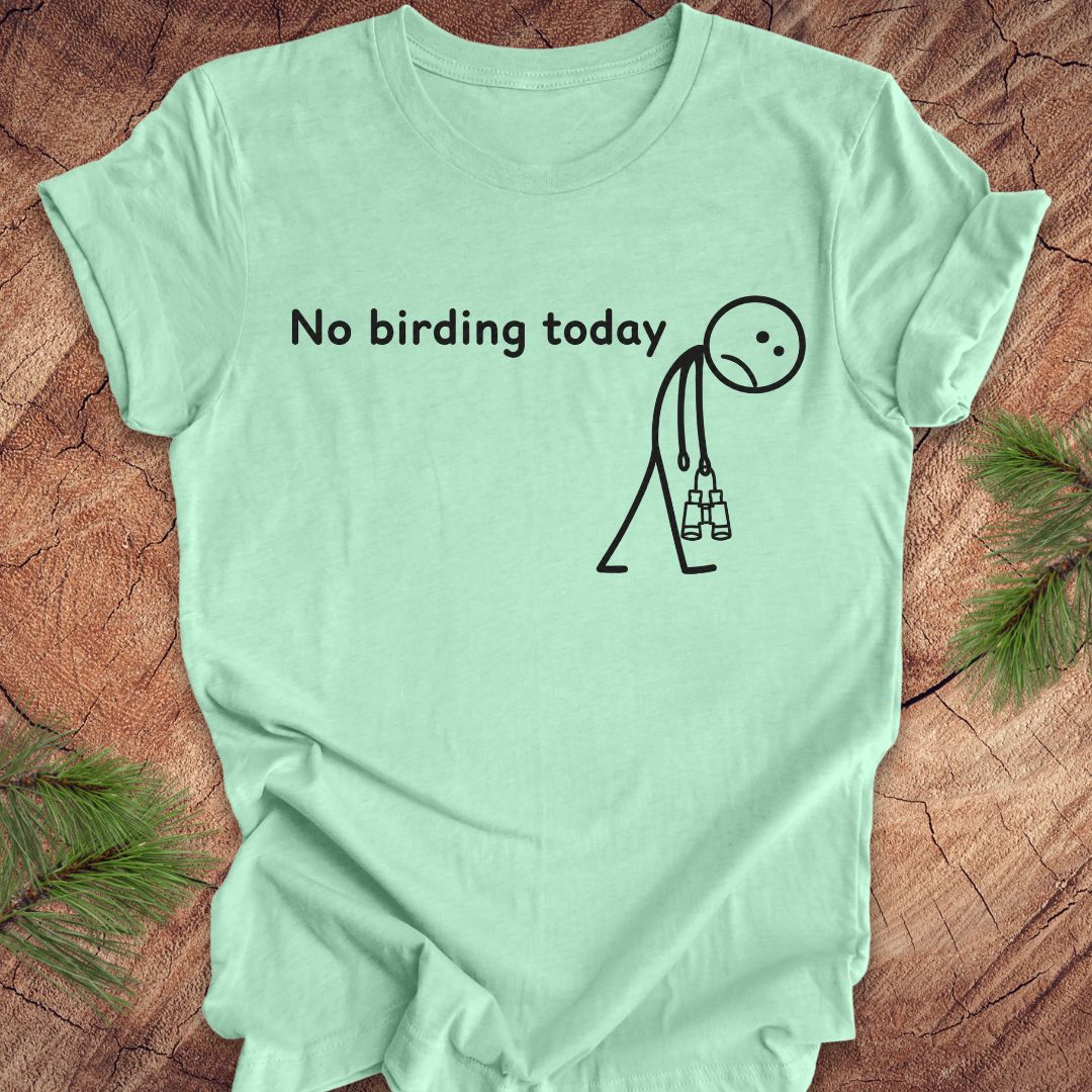 A mint green short-sleeve t-shirt with a black graphic print that reads 'No birding today' next to a stick figure holding binoculars, laid flat on a rustic wood background with pine needles in the corners.