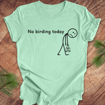 A mint green short-sleeve t-shirt with a black graphic print that reads 'No birding today' next to a stick figure holding binoculars, laid flat on a rustic wood background with pine needles in the corners.