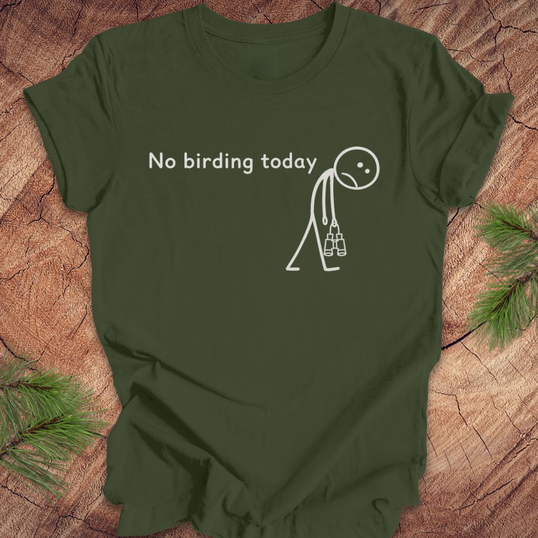 An olive green short-sleeve t-shirt with a white graphic print that reads 'No birding today' above a simple stick figure holding binoculars, laid flat on a rustic wood surface with pine branches in the corners.