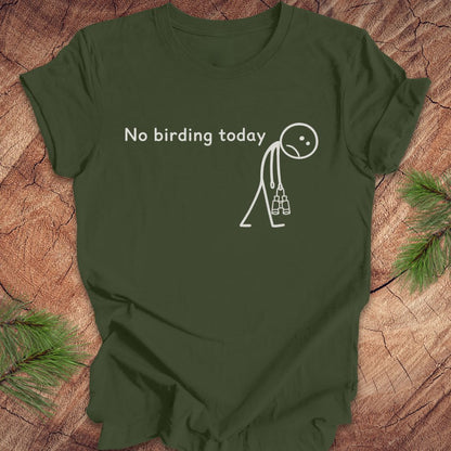 An olive green short-sleeve t-shirt with a white graphic print that reads 'No birding today' above a simple stick figure holding binoculars, laid flat on a rustic wood surface with pine branches in the corners.