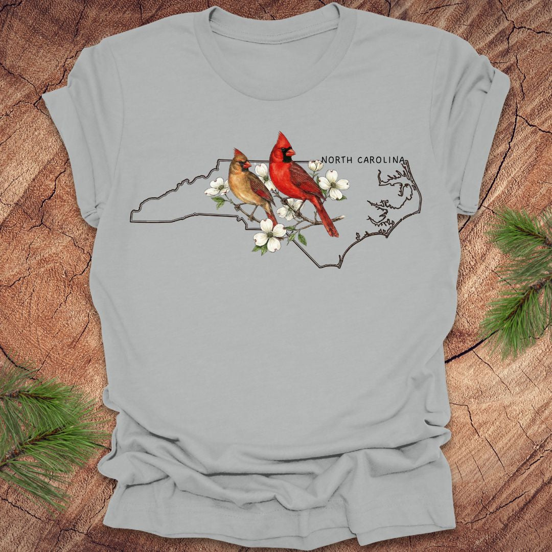 Gray t-shirt with a design of two cardinals on a branch with flowers and 'North Carolina' text, placed on a wooden surface.