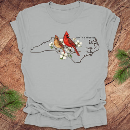 Gray t-shirt with a design of two cardinals on a branch with flowers and 'North Carolina' text, placed on a wooden surface.
