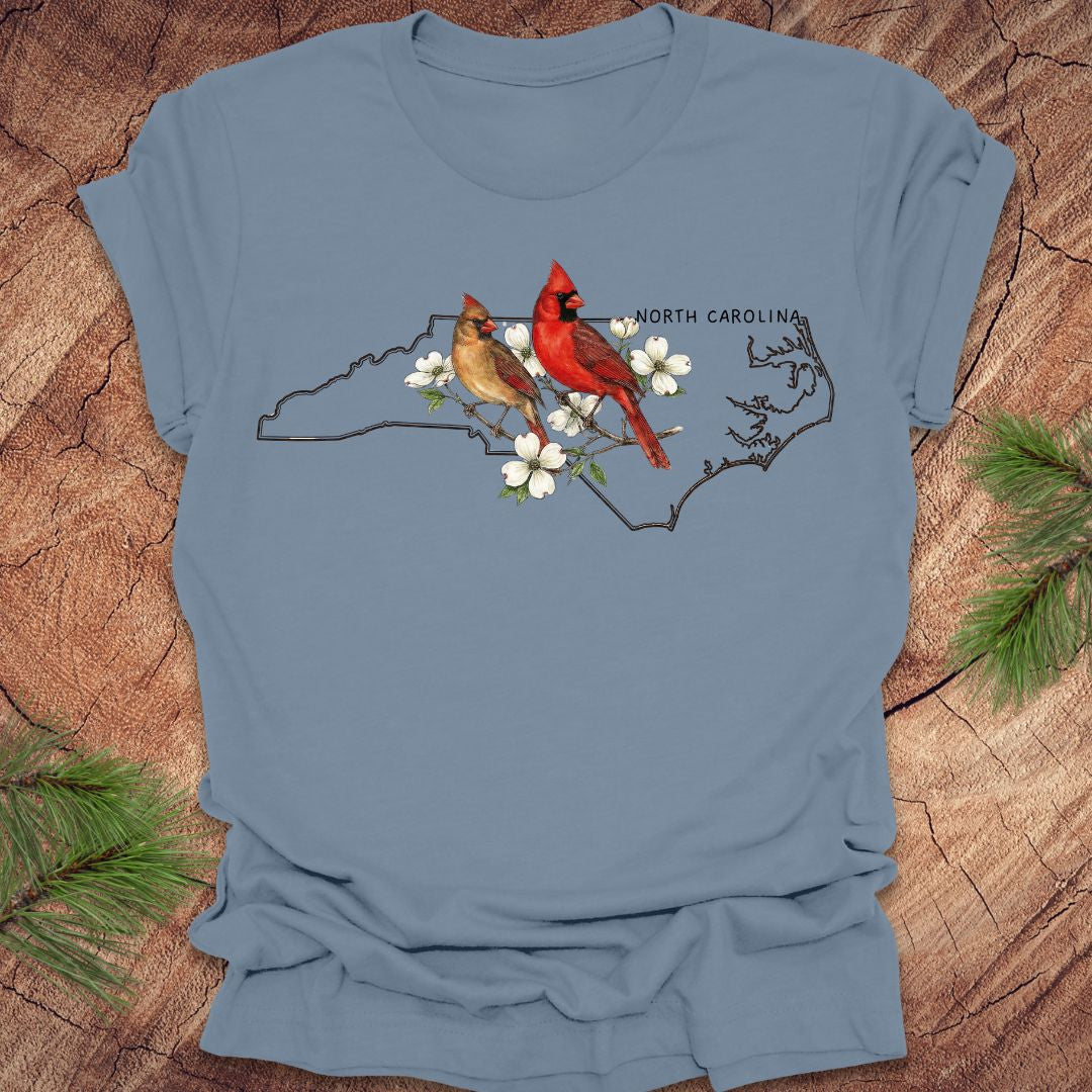 Blue t-shirt with a design of two cardinals on a branch with white flowers and 'North Carolina' text, placed on a wooden surface.