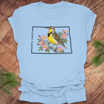 Baby blue t-shirt with a meadowlark and prairie rose flower inside the outline of North Dakota on a wooden surface