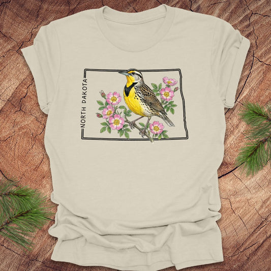 Beige t-shirt with a meadowlark and prairie rose flower inside the outline of North Dakota on a wooden surface